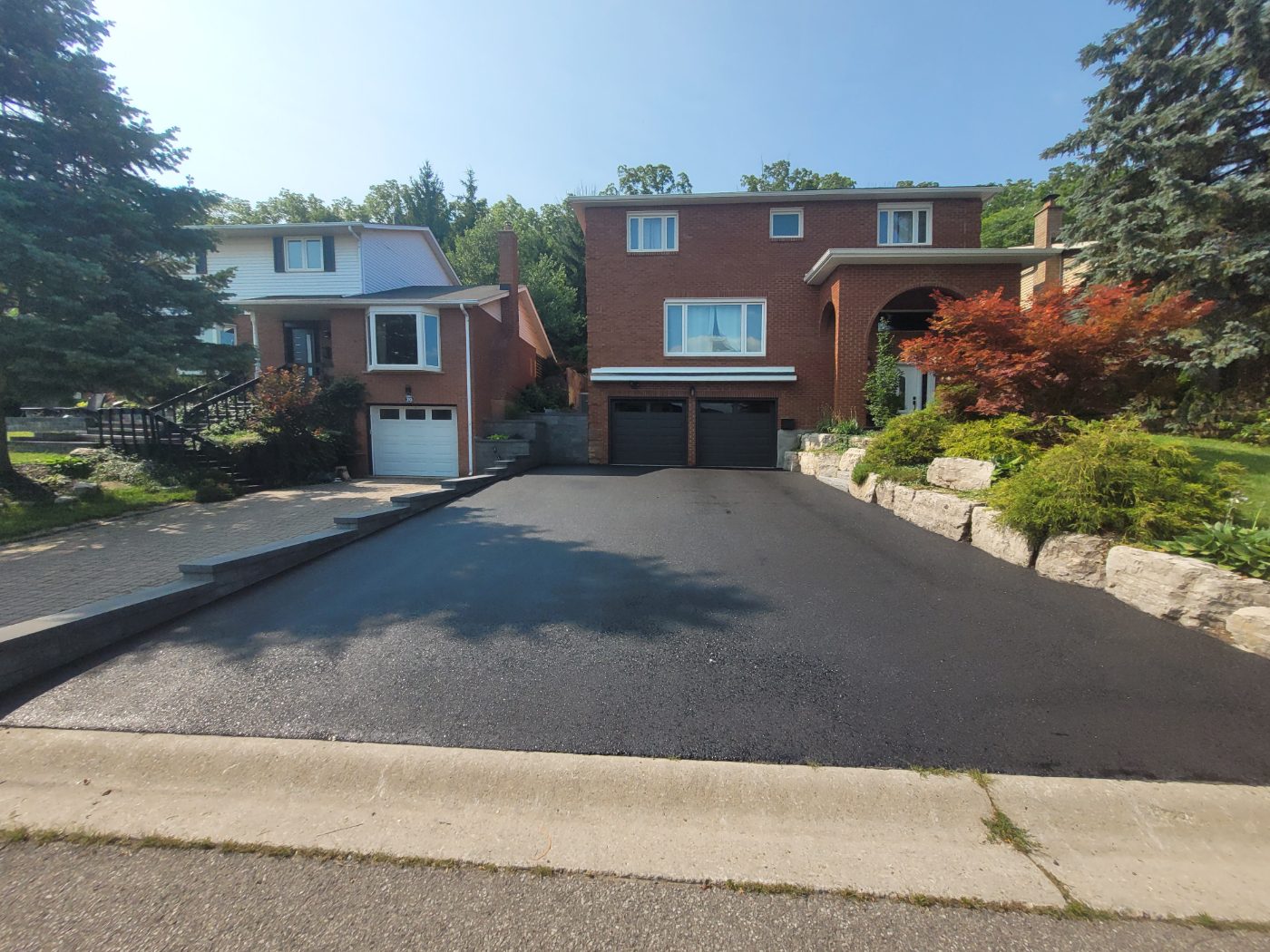 Wide double driveway freshly sealed between two brick homes with an armour stone garden wall